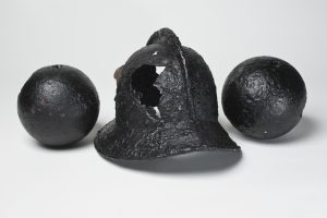 A black, historic helmet with a hole in it, and two black cannonballs