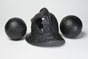 A black, historic helmet with a hole in it, and two black cannonballs