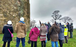 A group of about ten people, dressed in outdoor clothing and hard hats, stand on grass outside an old stone building (Clackmannan Tower), looking up.