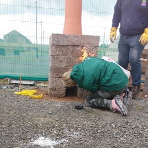 A person kneeling down, looking into a small kiln with flames shooting out