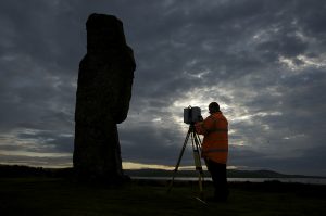 A person standing with a laser scanner pointing at a historic standing stone in the countryside