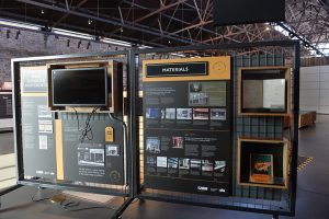 A black and yellow exhibition panel, with photos, wording and a screen on it