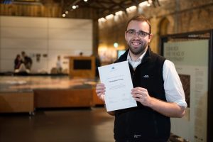 Chris standing inside the Engine Shed main space, smiling, and holding up a certificate