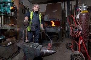 A blacksmith standing in a workshop in front of an anvil holding a hammer and piece of hot metal