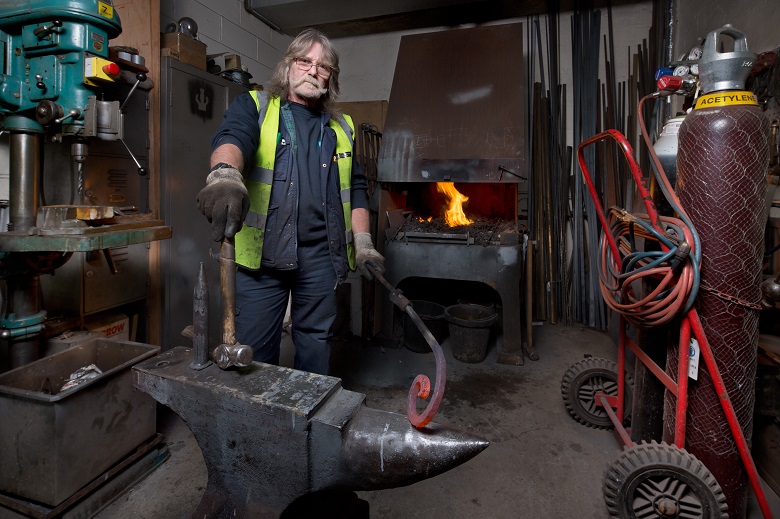 A blacksmith standing beside an anvil, holding a hammer and piece of hot metal