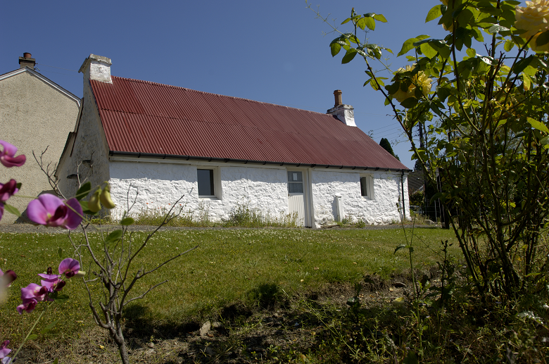 A historic cottage surrounded by a beautiful garden. The cottage has a corrugated iron roof