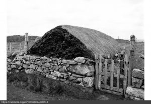 A black and white photo of an old thatched house surrounded by a stone rubble wall