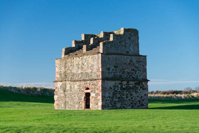 A large, square free-standing dovecoat surrounded by grass, with a stepped roof.
