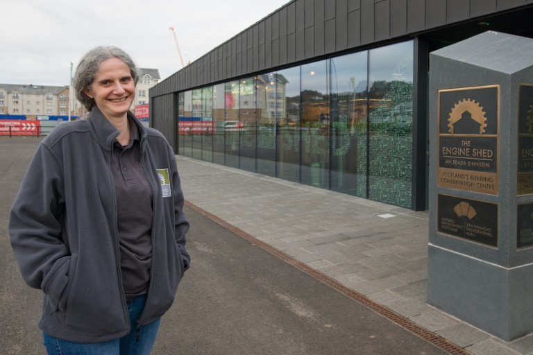 A person wearing a grey jumper, standing outside the glass-fronted Engine Shed building