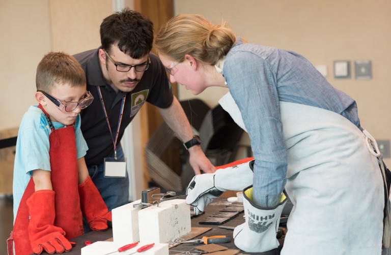 A person showing an adult and a child how to cast decorative items using metal. A piece of wood is place in between a clamp on the table.