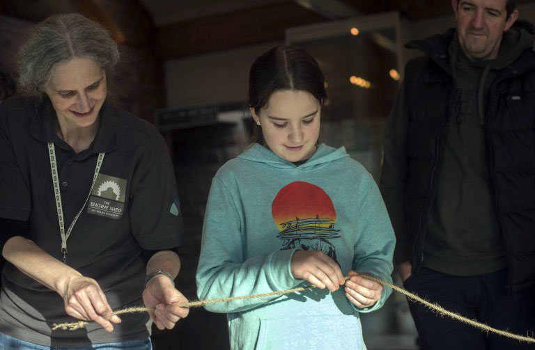 A person wearing a grey Engine Shed t-shirt, twisting a piece of rope with a young teenage child.