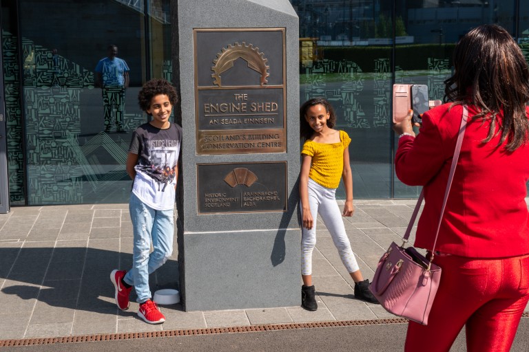 Two children having their photo taken by an adult beside the Engine Shed monolith