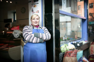 A shopkeeper in an apron, standing outside a shop entrance
