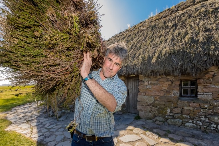A person holding a bundle of thatching material in front of a thatched house