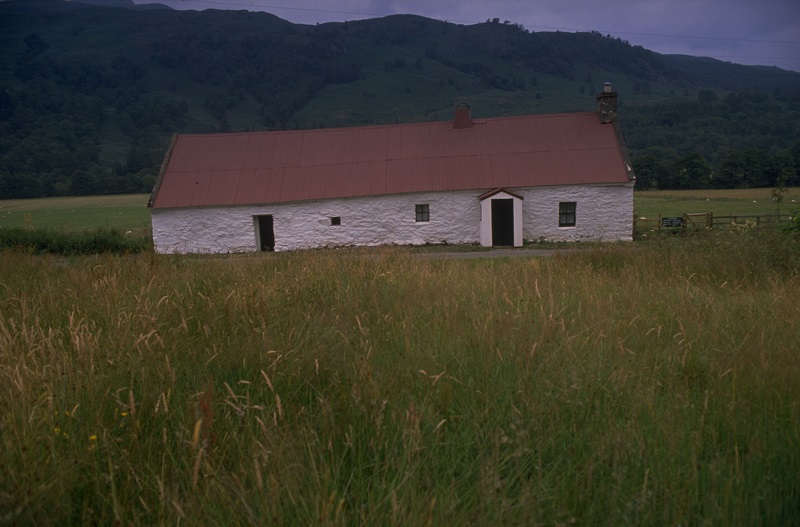 A white stone cottage with a corrugated iron roof, in a countryside setting with hills in the background