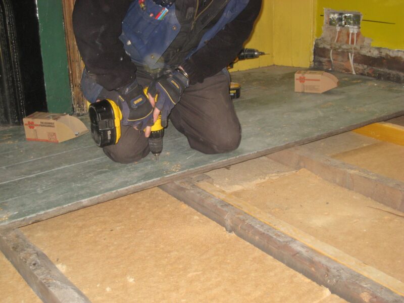 A person drilling into a wooden floorboard. Many of the other floorboards in the room have been lifted and removed