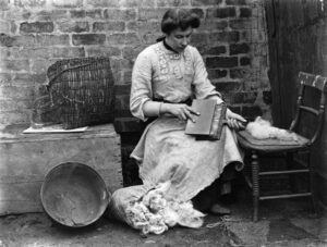 A woman wearing a long dress, sat at a chair, spinning wool onto the chair beside here