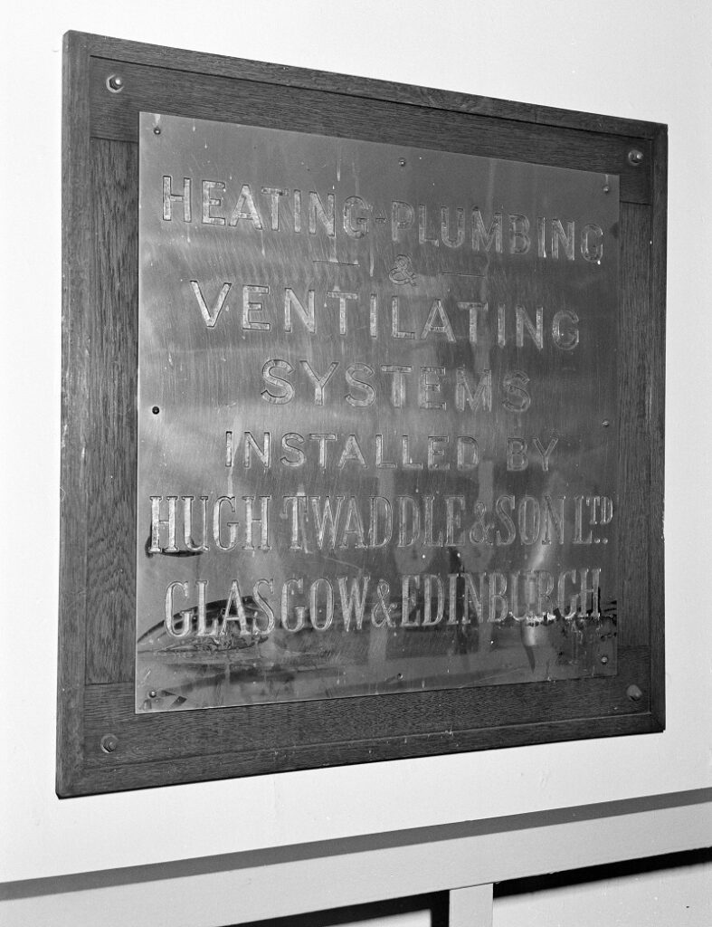 A metal plaque, on a wall, displaying the words "heating, plumbing, ventilation systems installed by Hugh Twaddle & Sons LTD, Glasgow and Edinburgh"