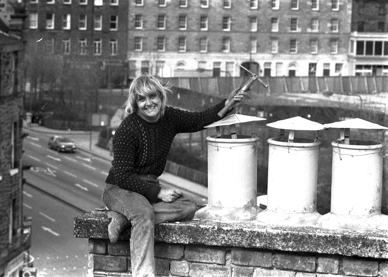 A woman slater, wearing a jumper and jeans, sitting on top of a chimney stack, holding a tool above the chimney pots