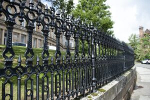 Black, highly decorative ironwork railings, running along the side of a pavement. They are fixed on top of a low, stone wall.