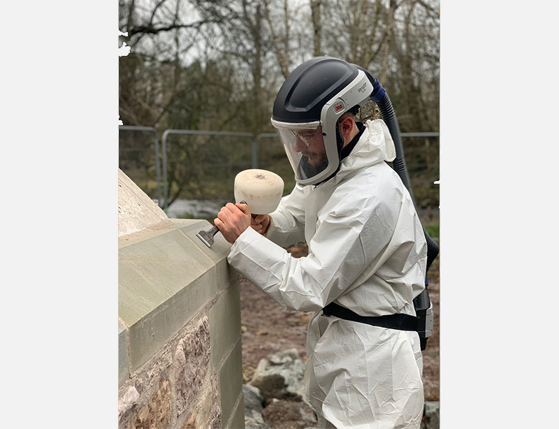 A person carving into a stone part of a bridge, wearing overalls and a helmet. They are using a chisel to carve.