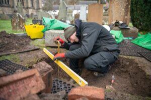 A person measuring a small trench dug out from the ground in a graveyard using a spirit level