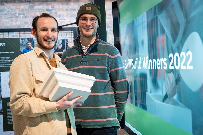 Two people smiling standing in front of a sign that reads 'SkillBuild Winners 2022'. One of them is holding a large piece of stone