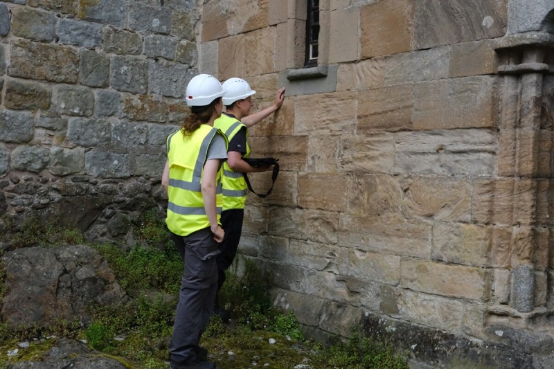 Two people wearing high vis vests and hard hats examining the stone wall of a historic castle.