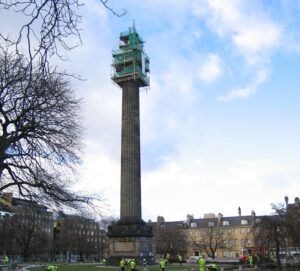 A tall, sandstone column monument in Edinburgh. There is scaffolding around the top of the monument and a group of workers wearing hard hats and high vis standing at the bottom © British Geological Survey