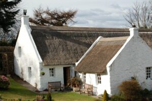 A traditional thatched building in Swanston. Looking at the front door, the t-shaped building is stonewashed white, with a sloping roof of dark thatch.