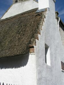 A close-up of a thatched roof of a building in Cromarty. The roof is sloped and made of tightly-packed dark thatch, the contrasting building is whitewashed stone, with the sun shining behind.
