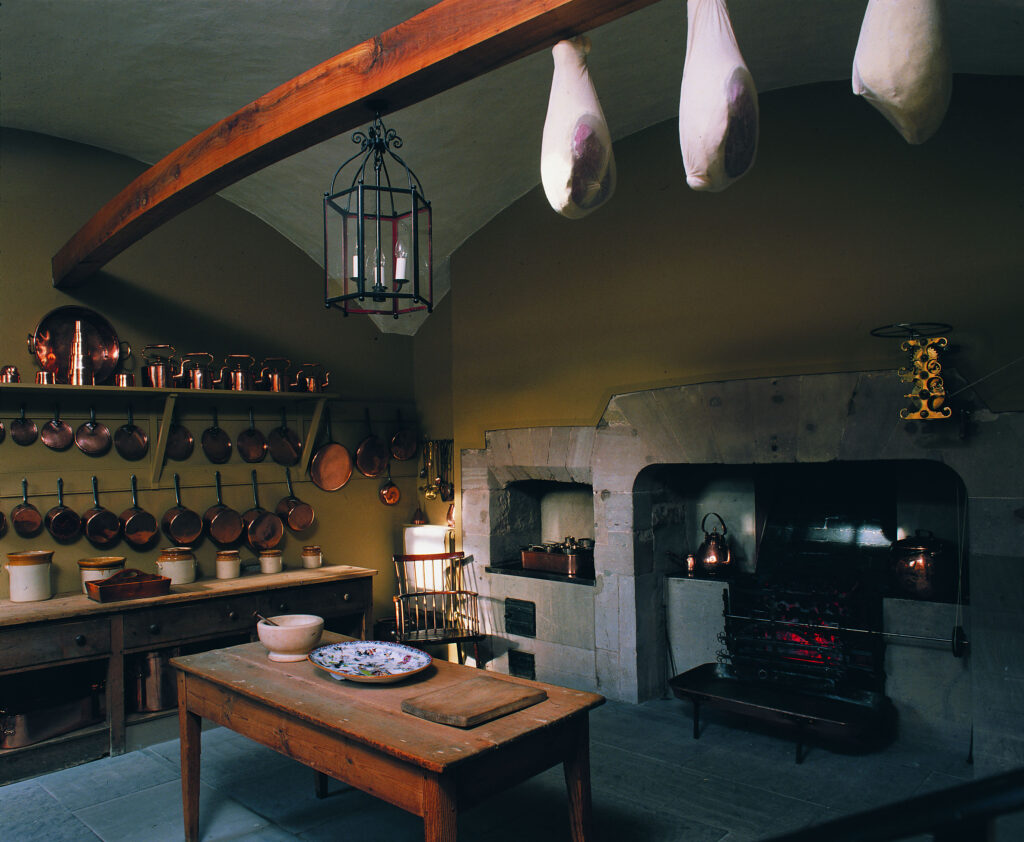 A historical kitchen, set in a grand old house. There is a large range fire, and a selection of around thirty pots and pans hanging from the wall. A table in the centre has bowls and plates on it, and three hams hang from the rafters.