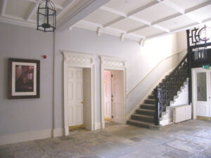 A light grey coloured hallway with a grey stone floor. There is a painting on the wall and to the right, a large staircase with a wooden banister.