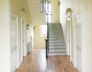 A hallway and stairs in an old period house. The walls and doors are painted white and the floor is lightly varnished brown. The stairs go straight up to a landing with a picture window. A large black light fitting hangs from the ceiling.