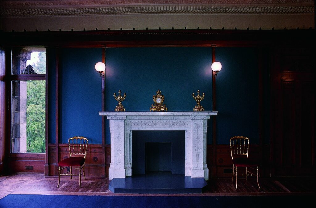 A marble fireplace in front of a deep blue wall in an old building. There are two globe lamps and a pair of wooden chairs, one either side of the fireplace. To the left is a window, looking out onto trees.