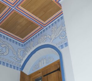 A section of restored painted ceiling in Stirling Palace. The image shows a panelled wooden ceiling and a curving blue-lined doorframe, with in between a colourful blue fresco of heraldic lions.