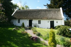Image shows a historical thatched cottage on the outskirts of Edinburgh. The cottage is painted white, with two doors and two windows and a long sloping roof of grey thatch. In the foreground is a well tended garden with grass, plants and flowers.