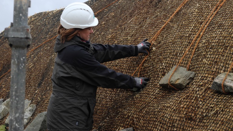 A person wearing a hard hat, pulling on a rope and large stone that is weighing down netting on top of a thatched roof