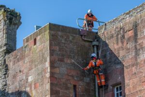 One person looking over the top of a building at another person who is hanging from the site of a building with a rope, tapping the stonework with a large stick. Both are wearing high-vis overalls and hard hats.