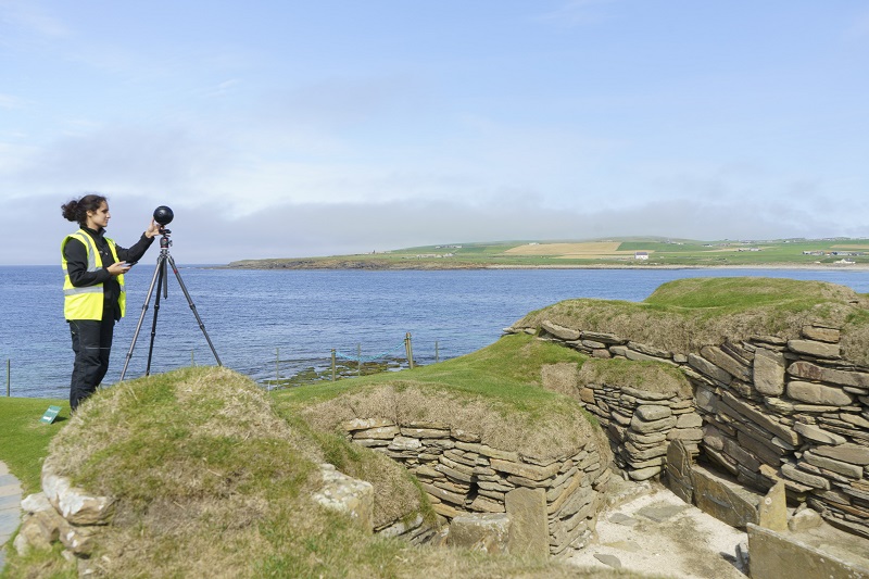 A person wearing a yellow high-vis vest, standing, looking into a laser scanner at Skara Brae. Skara Brae is made up of low stone walls topped with turf, by the sea.