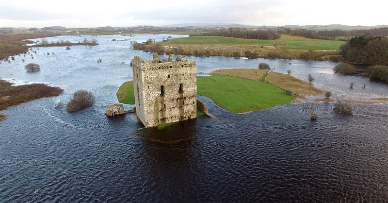 A small, rectangular historic castle. The surrounding farmland has flooded, and water completely surrounds the castle.