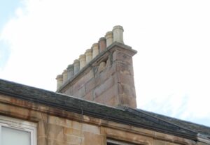 A roof of a traditional building seen in close-up, with the chimney stack showing visible long term weather damage. There are holes in the masonry.
