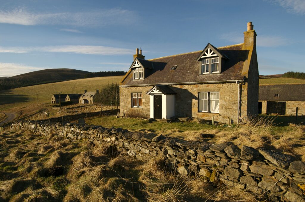 A traditional cottage, with two windows on the ground floor and two dormer windows in the roof. There are other cottages in the background surrounded by farmland.