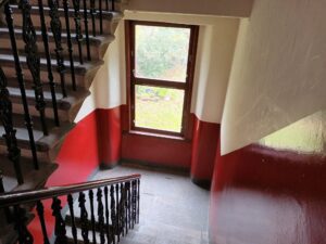 A stairwell in a historic tenement, showing ornate balustrates and a two tone painted stair, which is dark red and cream in colour. There is a window overlooking a garden.