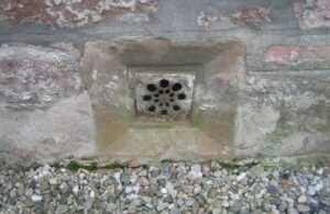 A circular series of small holes at the base of a wall, the wall vent sits in the grey brickwork of a building, with a gravel path beneath.