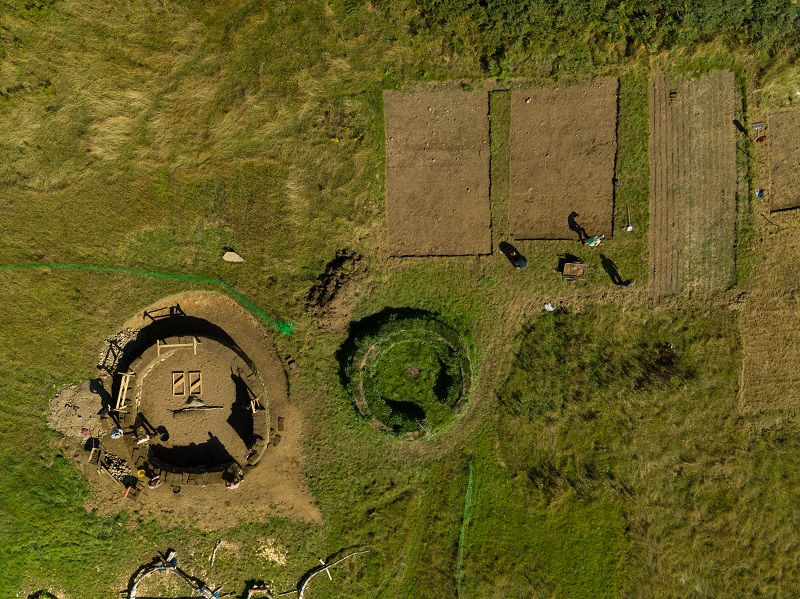 A birds-eye view of a building site for a hut. The walls of the turf hut are under construction, and nearby there are bare patches of ground where turf has been cut to build the hut