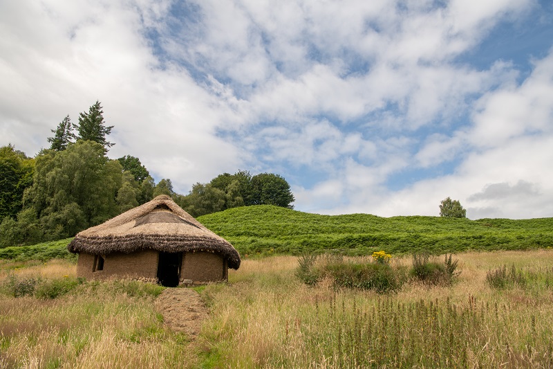 A small, thatched roof hut in the countyside. The hut is made from earth and has a large, rectangular opening for a door.