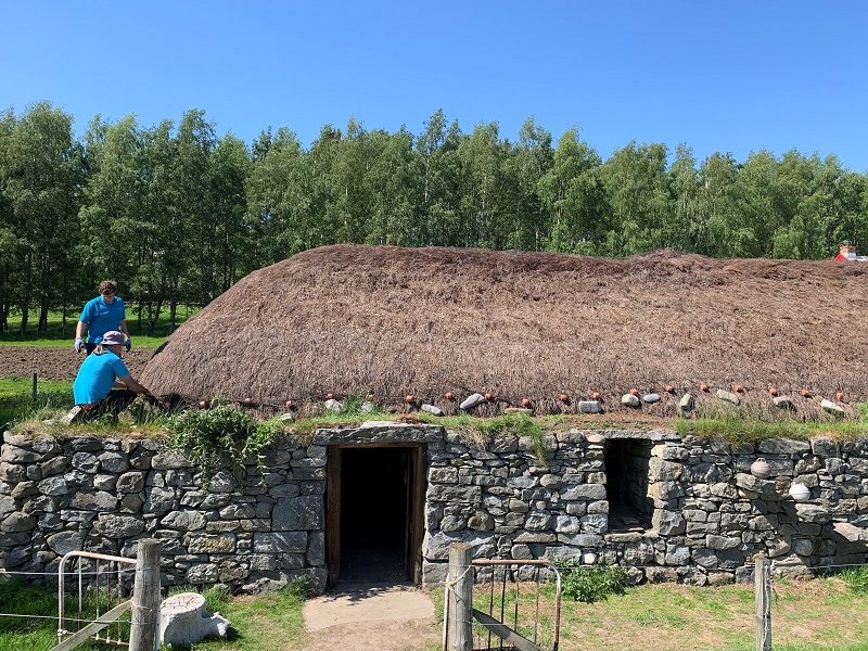 A thatched single-storey building made from stone. Two people are inspecting the thatch roof.