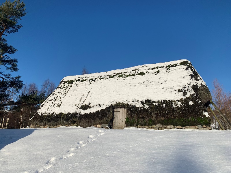 An old building with a long, thatched roof and a small, wooden door. The roof and ground is covered with snow.