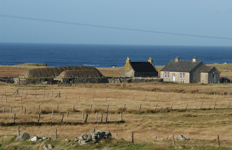 Two low-to-the-ground thatched buildings, with roofs netted down with stones, and two traditional stone buildings with chimneys and slate roofs. The buildings are surrounded by fields and the sea is behind them.